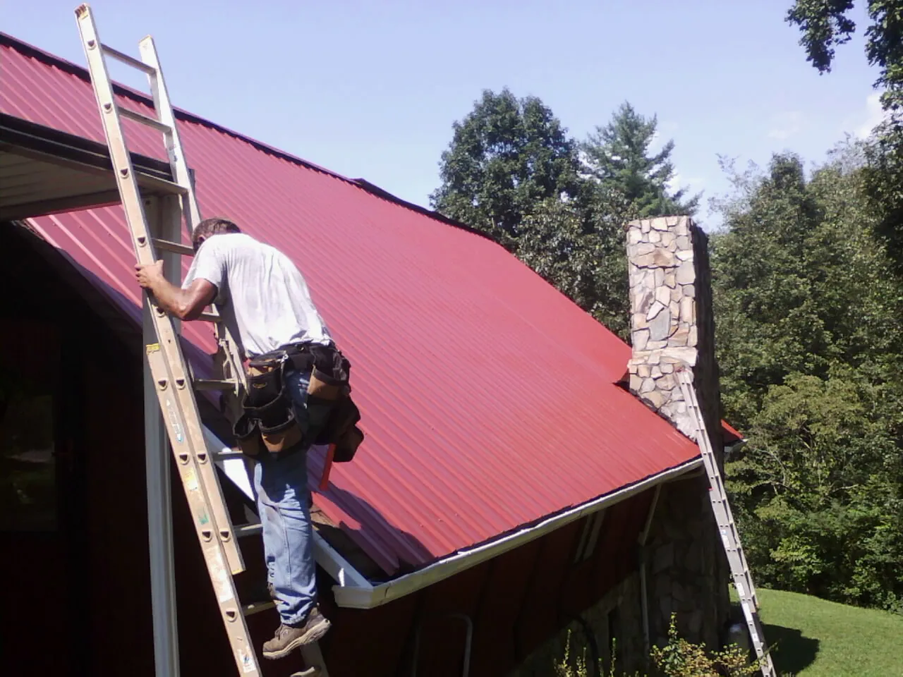 Roofer on a ladder beside a red metal roof and stone chimney — Soffit & Fascia Repair by Roofing Brothers in Londonderry